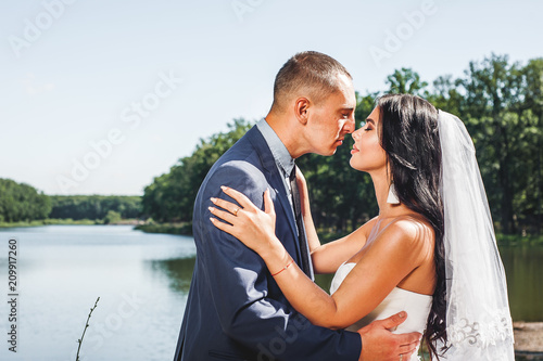 The bride and groom stand by the river, smiling and kissing