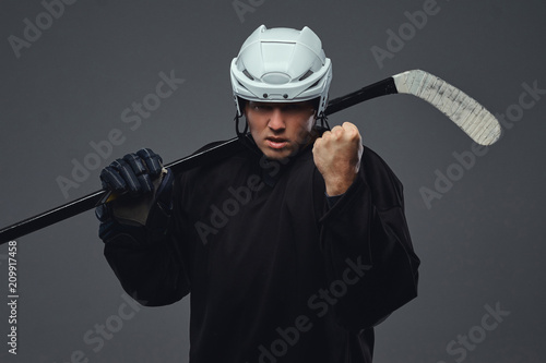 Photography Angry professional hockey player in black sportswear standing with a hockey stick on a gray background