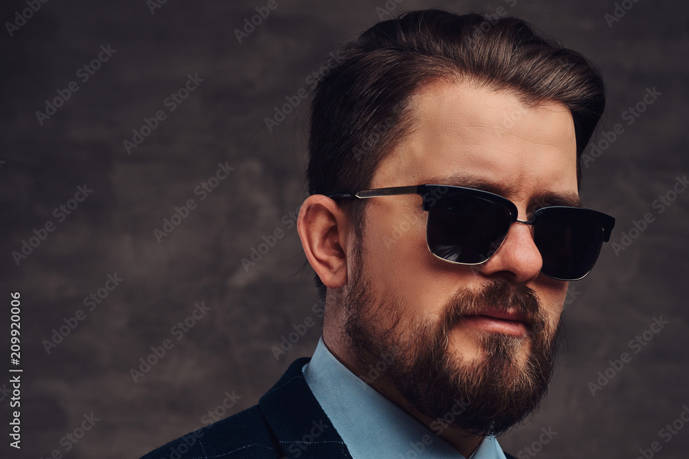 Close-up portrait of a handsome fashionable middle-aged man with beard and hairstyle dressed in an elegant formal suit and sunglasses. Isolated on a textured dark background in studio. Close-up portrait of a handsome fashionable middle-aged man with beard and hairstyle dressed in an elegant formal suit and sunglasses. Isolated on a textured dark background in studio.
