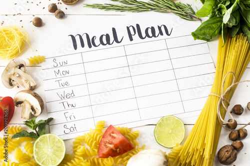 A meal plan for a week on a white table among products for cooking - pastas, basil, vegetables, lime, seeds, nuts and spices. Top view, flat lay, copyspace