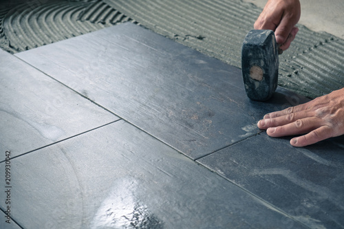 Worker placing ceramic floor tiles on adhesive surface, leveling with rubber hammer