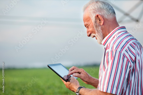 Happy and satisfied gray haired senior agronomist or farmer using a tablet in soybean field. Irrigation system in background. Side view.