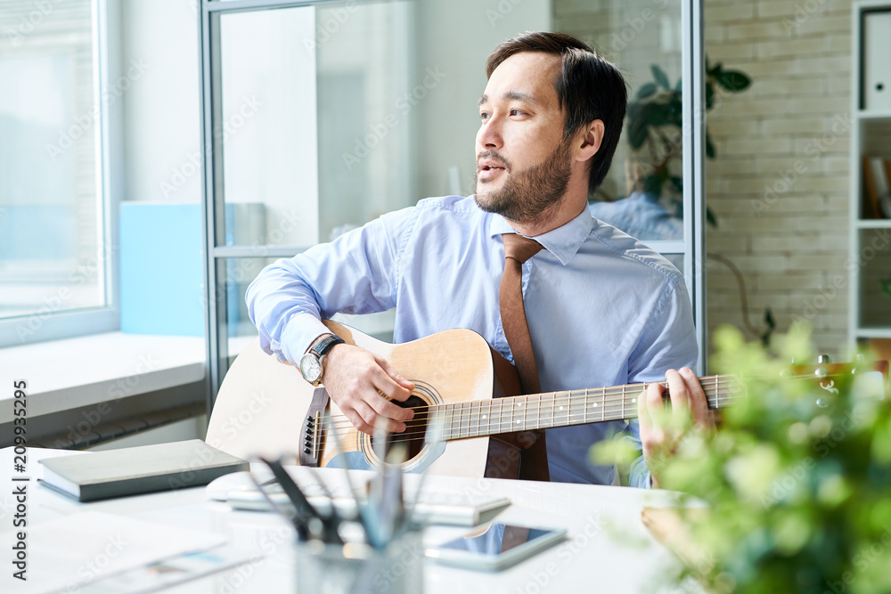 Bearded man in formal outfit sitting at desk in office and chilling with guitar playing melody. Bearded man in formal outfit sitting at desk in office and chilling with guitar playing melody.