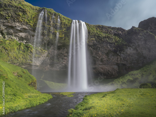 Seljalandsfoss - island - Wasserfall von einer klippe sich herabst  rzend in einen See vor Felswand bei Blauem Himmel und Sonnensschein