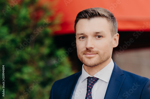 Headshot of attracttive successful male manager has confident serious expression, looks at camera, dressed in formal clothes, poses outside, going to have business meeting with partners abroad. photo