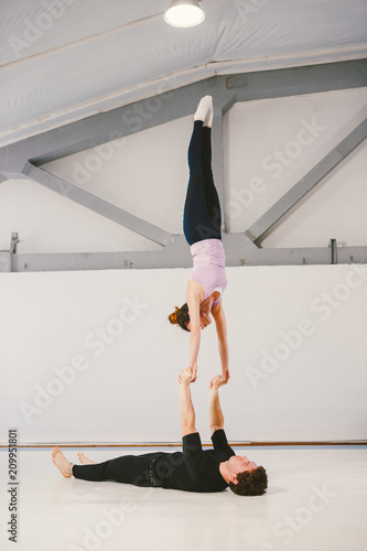 young Caucasian male and female couple practicing acrobatic yoga in a white gym on mats. Loy HAND-TO-HAND, Skills Wind Mill, Position Type L-Bass, Difficulty Really Hard, Number of Persons 2 Person