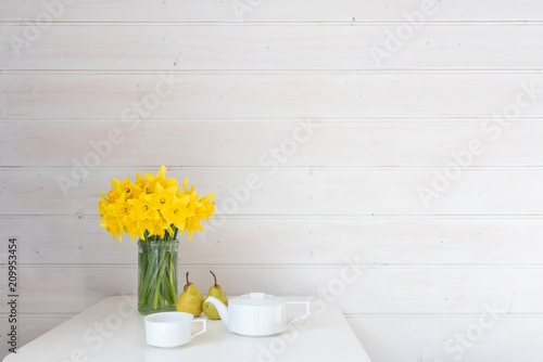 Fototapeta Naklejka Na Ścianę i Meble -  Yellow daffodils in glass jar on white table with two green pears and white teapot against rustic white wood panelled wall