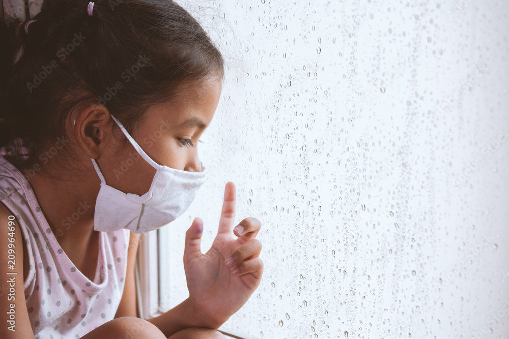 Sick asian little child girl wear protective mask looking outside through the window in the rainy day Sick asian little child girl wear protective mask looking outside through the window in the rainy day