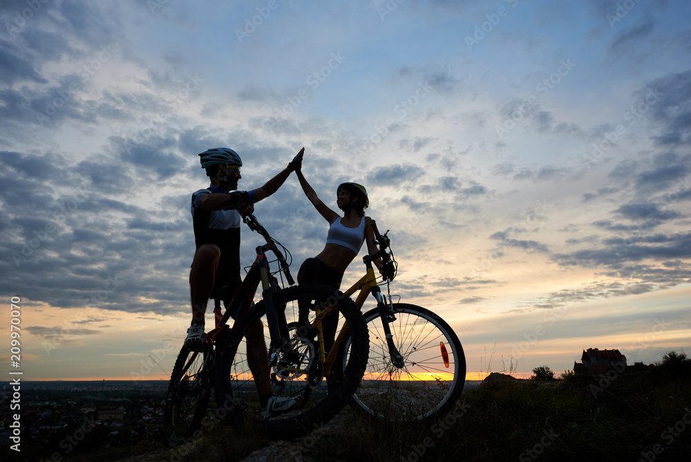 Obraz premium Athletic man and sttractive woman in helmets highing five and looking at each other after cycling. Sporty couple of cyclists sitting on bikes and resting in twilight. Evenening sky background.