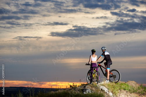 Fototapeta Naklejka Na Ścianę i Meble -  Back view of a couple riding a mountain bike on top enjoying the sunset and a beautiful evening in the mountains. Young people dressed in sporting clothes and helmets