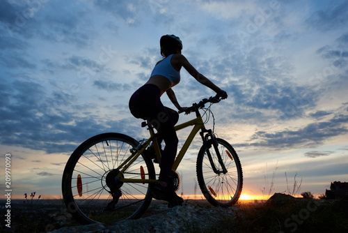 Back view of young woman cycling mountain bicycle. Silhouette of female cyclist in helmet enjoys sunset on a top of mountain under fairytale sky with clouds and bright sun in the evening