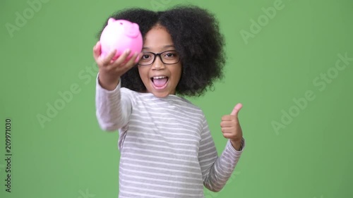 Young cute African girl with Afro hair holding piggy bank and giving thumbs up