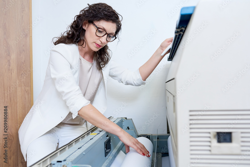 Elegant adult woman in glasses putting roll of paper into plotter while working in nice printing office. Elegant adult woman in glasses putting roll of paper into plotter while working in nice printing office.
