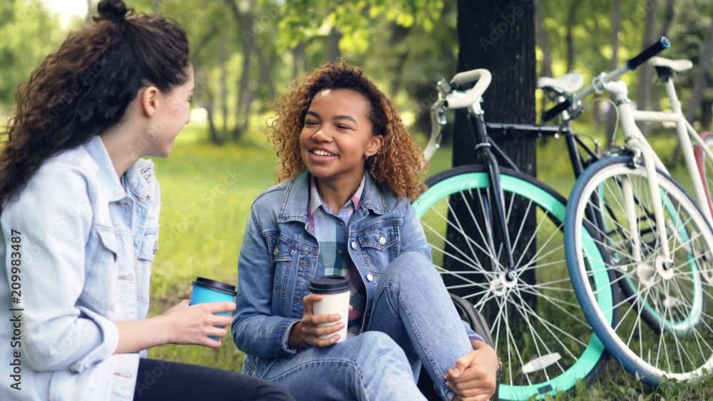 Young African American woman is chatting with her Caucasian friend and drinking takeaway coffee relaxing in beautiful park after riding bikes on sunny summer day. Young African American woman is chatting with her Caucasian friend and drinking takeaway coffee relaxing in beautiful park after riding bikes on sunny summer day.