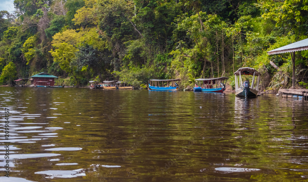 The beginning of the Tahan River after the confluence with the Tembeling River in Taman Negara National Park, Malaysia. A few motorized long-tail boats with metal roofs are tied up at the riverbank.