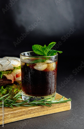 Glass of cola with ice, mint and rosemary on black background big sandwich, street food