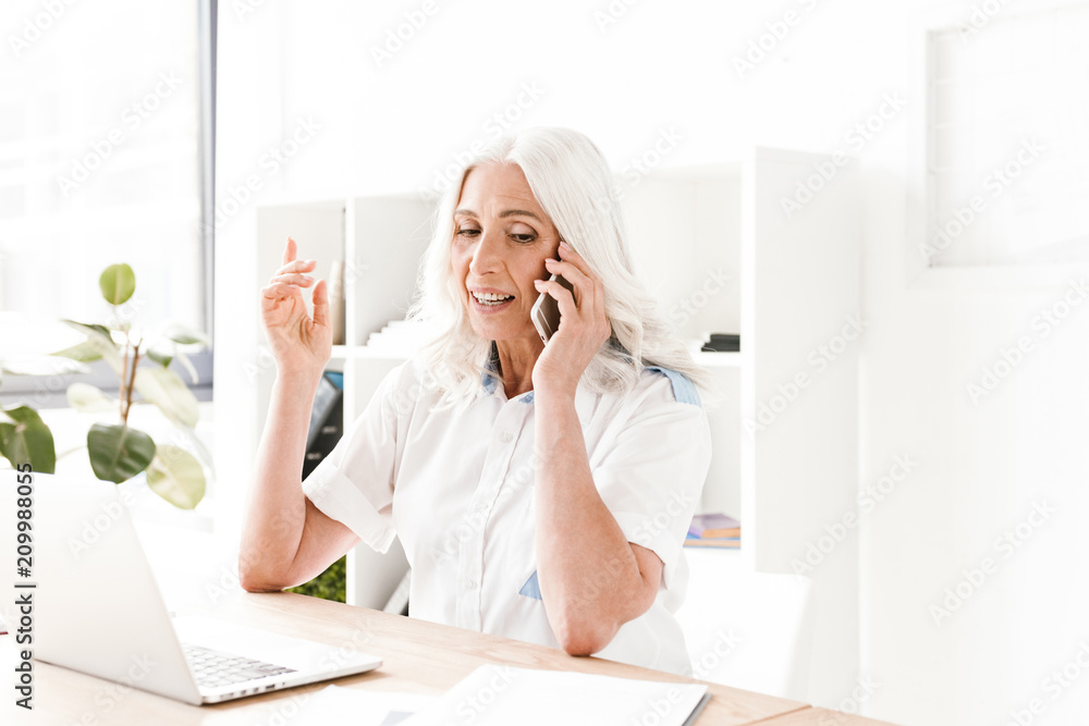 Mature woman talking by mobile phone using laptop computer.