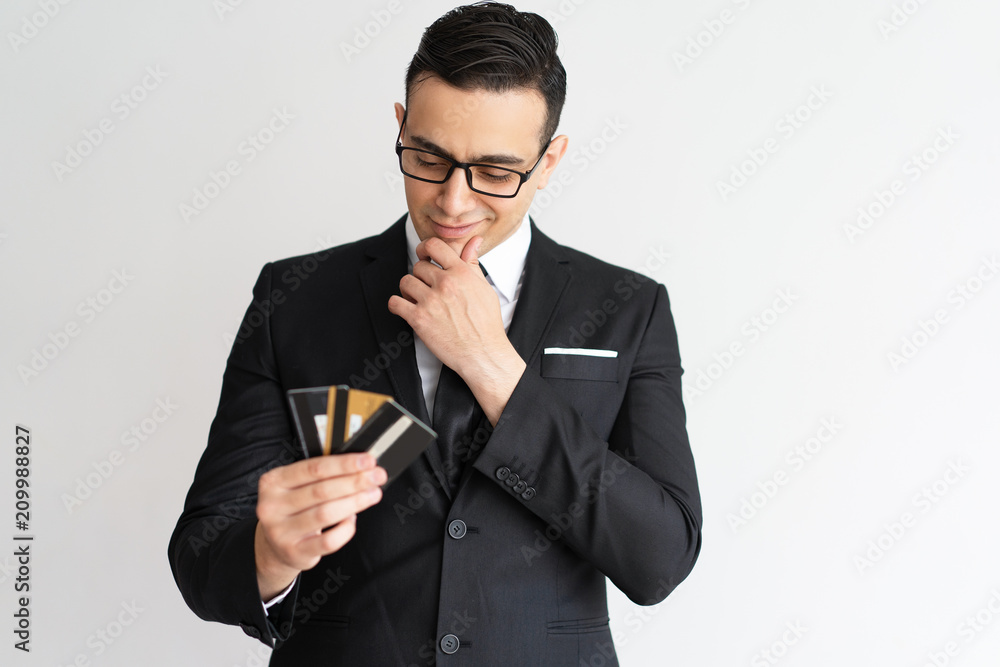 Thoughtful mixed race businessman looking at credit cards. Pensive handsome young male manager in glasses thinking of his profit and choosing credit card. Many opportunities for business concept Thoughtful mixed race businessman looking at credit cards. Pensive handsome young male manager in glasses thinking of his profit and choosing credit card. Many opportunities for business concept