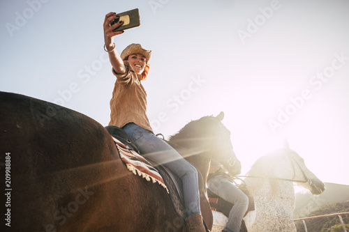 couple riding horses take a selfie with modern technology smartphone. cowboy lifestyle and smile woman. sunset time and backlight for outdoor leisure activity for friends
