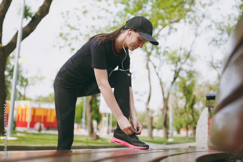 Young athletic beautiful brunette girl in black uniform and cap with headphones listening music, tying shoelaces before running, training on bench in city park outdoors. Fitness, healthy lifestyle