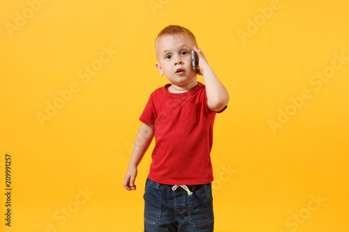 Little kid boy 3-4 years old in red t-shirt talking on mobile phone, conducting pleasant conversation isolated on yellow background. Kids childhood lifestyle concept. Problem of children and gadgets.