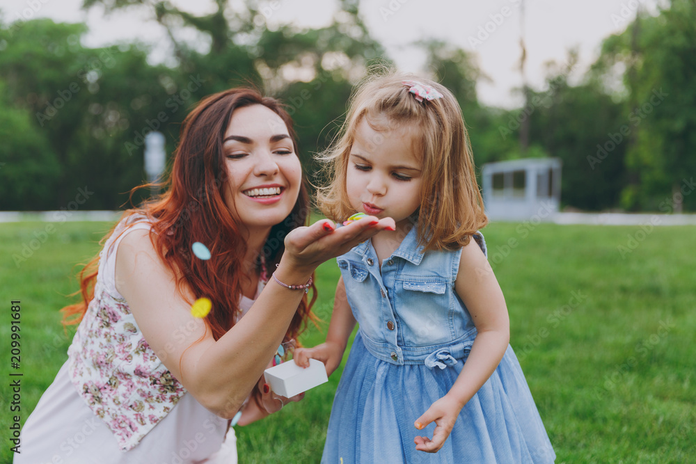 Fototapeta premium Woman in light dress and little cute child baby girl play and blowing on colorful paper confetti in green park. Mother, little kid daughter. Mother's Day, love family, parenthood, childhood concept.