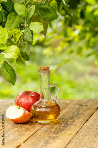 Apple vinegar in glass bottle and fresh red apples on wooden boards with green natural background