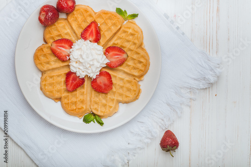  Norwegian heart shaped waffles topped with strawberries,  mint and whipped cream on white plate and white wooden background. St. Valentine's Day breakfast. Romantic breakfast. Copy space