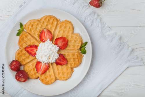  Norwegian heart shaped waffles topped with strawberries,  mint and whipped cream on white plate and white wooden background. St. Valentine's Day breakfast. Romantic breakfast. Copy space