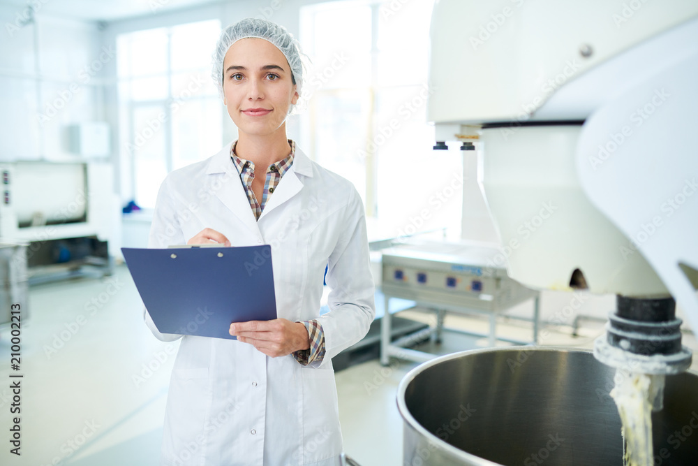 Confectionery factory employee standing in white coat near operating machinery holding clipboard and looking at camera. 