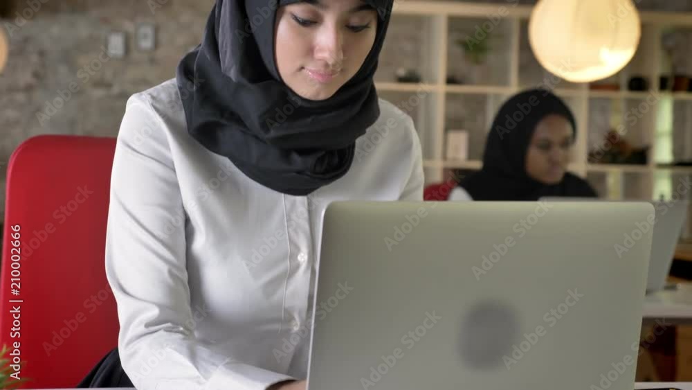 Portrait of young muslim women in hijab working and typing on laptop, concentrated, two womens sitting in modern office Portrait of young muslim women in hijab working and typing on laptop, concentrated, two womens sitting in modern office
