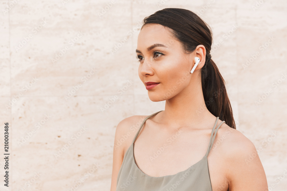Portrait of beautiful brunette woman wearing casual summer clothing and bluetooth earpod looking aside, while standing against beige wall outdoor