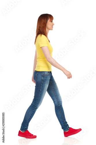 Young Woman In Yellow Shirt, Jeans And Red Sneakers Is Walking And Looking Away