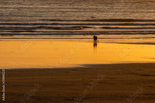 Couple walking their dog on the beach at sunset. Surfers silhouettes in the distance