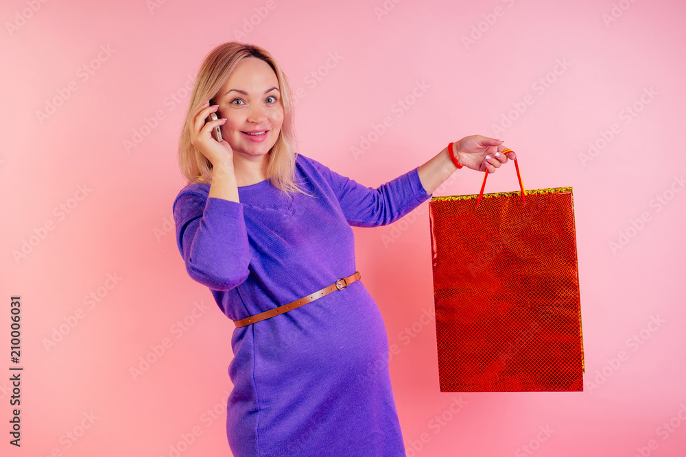 Beautiful blonde smiley pregnant woman big baby bump holding shopping bags and talking on the phone in the studio on a pink background . buying clothes for pregnant concept Beautiful blonde smiley pregnant woman big baby bump holding shopping bags and talking on the phone in the studio on a pink background . buying clothes for pregnant concept