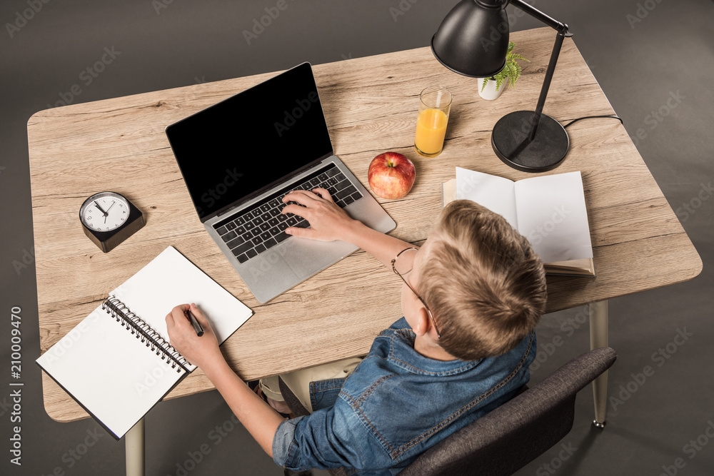 elevated view of schoolboy doing homework on laptop at table with textbook, book, plant, lamp, clock, apple and glass of juice elevated view of schoolboy doing homework on laptop at table with textbook, book, plant, lamp, clock, apple and glass of juice