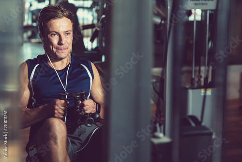 Concentrated young sportsman using rowing machine and listening to music in headphones while having intensive training at gym, portrait shot