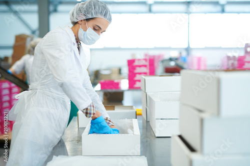 Fotografija Side view of confectionery factory worker in white coat putting baking parchment into empty paper box