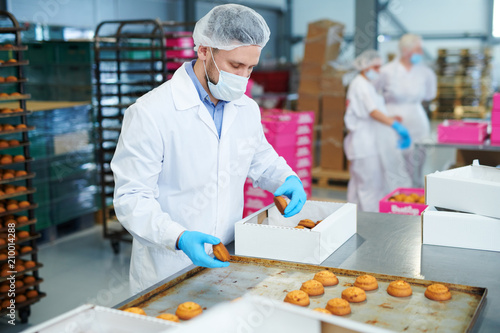 Confectionery factory worker in white coat collecting freshly baked pastry from tray and putting it into paper box.  photo