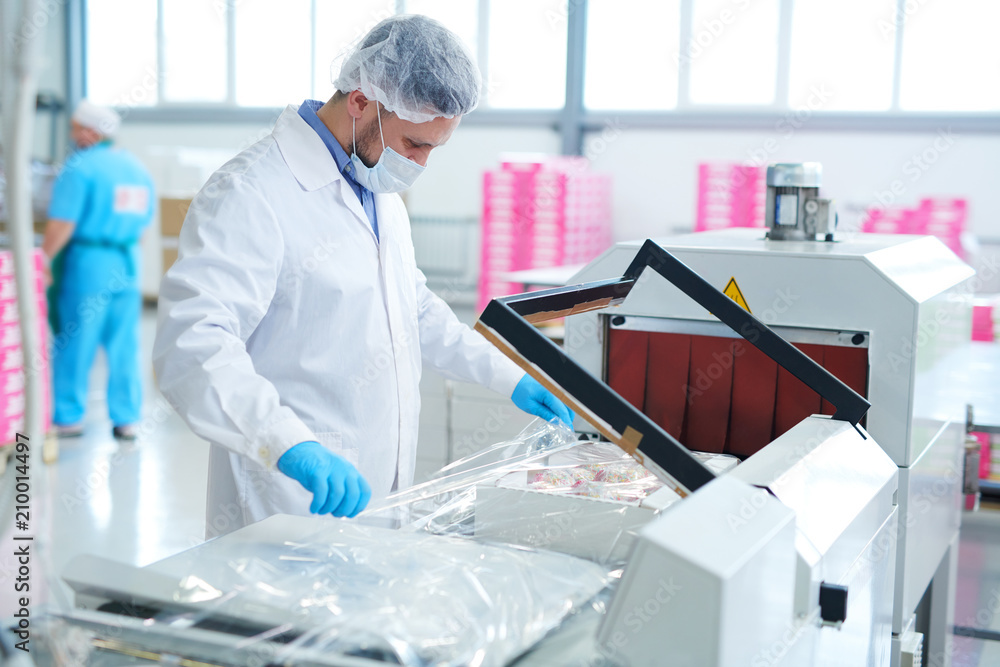 Confectionery factory employee in white coat wrapping paper box with pastry into packaging film.