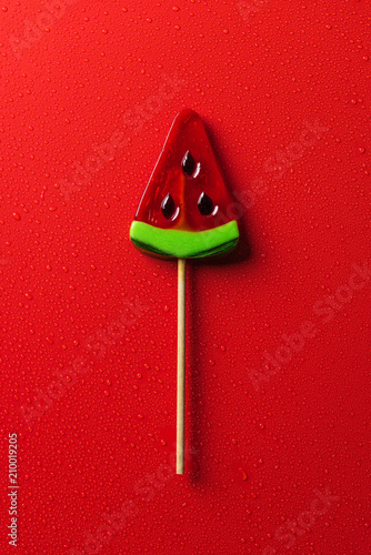 top view of lollipop in shape of watermelon on red surface with water drops