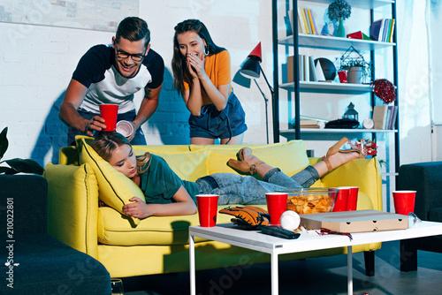 laughing young people looking at drunk female friend sleeping on sofa after home party