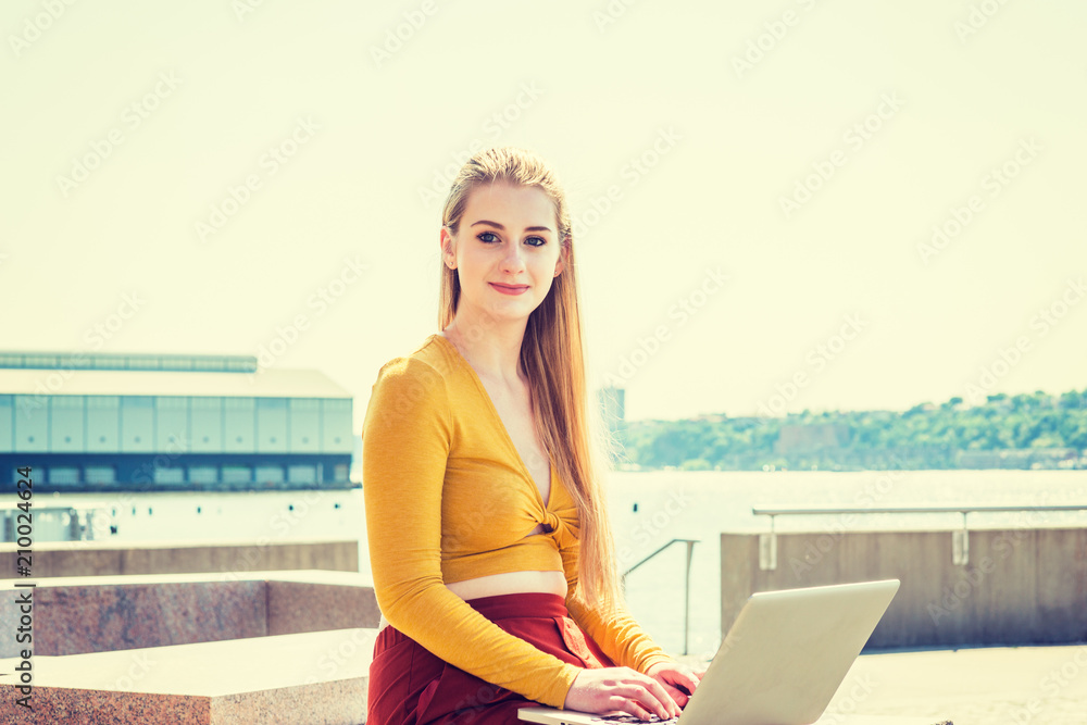 Young American teenage college student studying in New York, wearing yellow long sleeve deep v neck fit crop top, sitting by Hudson River under sun, working on laptop computer, looking, thinking..