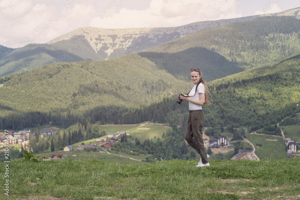 Girl with a displeased face and camera standing on a background of mountains. Forest on background