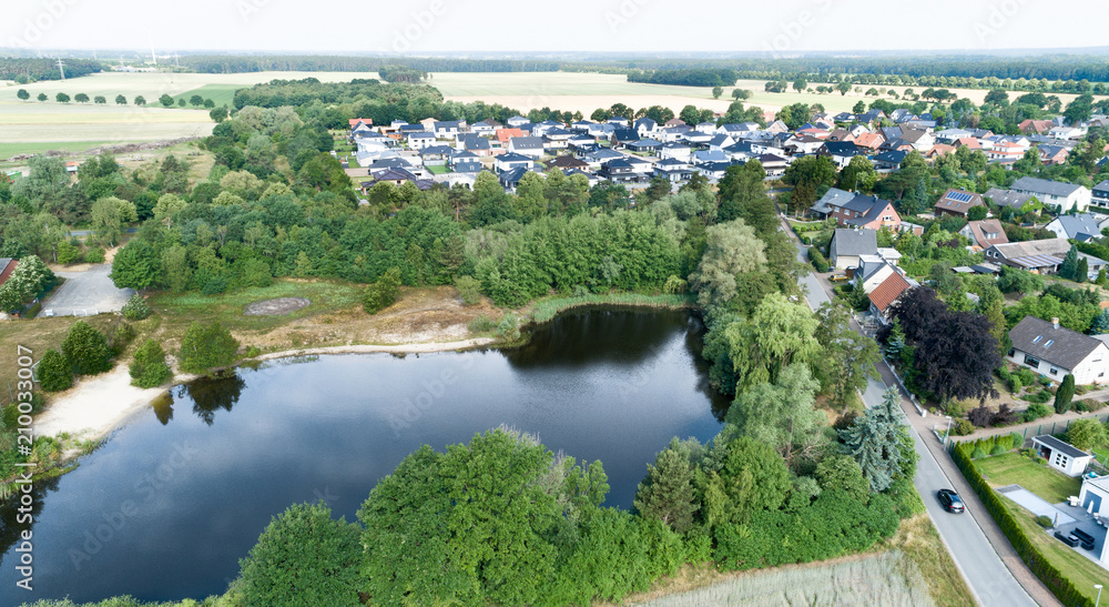 Fototapeta premium Aerial view of a suburb behind a small lake and a wooded area, with detached houses, semi-detached houses and terraced houses with small front gardens and green lawns in northern Germany