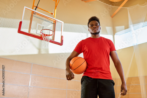 Handsome African-American man holding ball and looking at camera while standing on basketball court in gym.  © Seventyfour