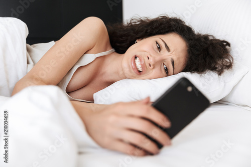 Image of frustrated upset woman 20s with dark curly hair lying in bed on white pillow after sleep, and looking at mobile phone with confusion