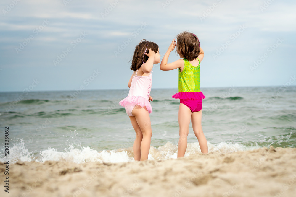 two little girls sisters are standing and playing on the sand at the beach on a summer day two little girls sisters are standing and playing on the sand at the beach on a summer day