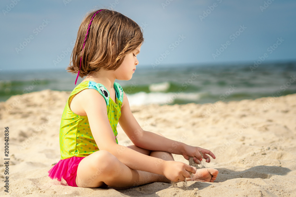 little girl in green swimsuit sitting on the sand at the beach on a summer day little girl in green swimsuit sitting on the sand at the beach on a summer day