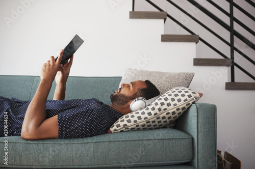 Man Lying On Sofa At Home Wearing Headphones And Watching Movie On Digital Tablet